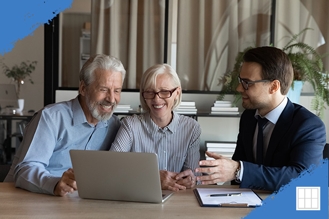 A loan officer in a suit discusses home loan approval with an older couple, smiling and looking at a laptop in a modern office setting with plants and bookshelves in the background.