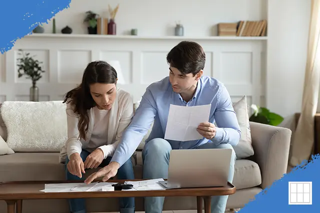 A young couple sitting on a couch reviewing mortgage documents and floor plans on a coffee table with a laptop, discussing home buying options.