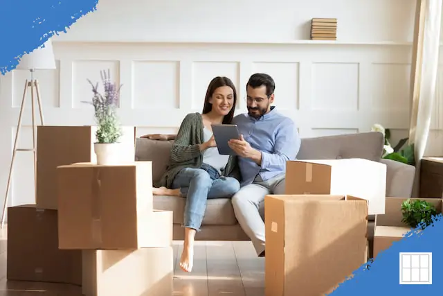 A smiling young couple sits on a couch in their new home, surrounded by moving boxes, as they look at a tablet together. The bright, modern living room features white walls, wooden floors, and potted plants.