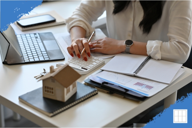 A woman calculating savings on a refinance using a calculator, miniature house, and financial reports, representing home equity in Overland Park.