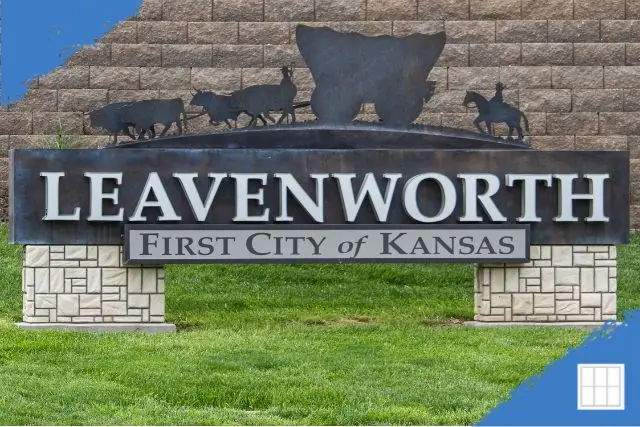 A metal welcome sign for Leavenworth, Kansas, reading "LEAVENWORTH First City of Kansas" with silhouettes of oxen pulling a wagon and a horseback rider atop a curved panel, set against a brick wall and grassy base.